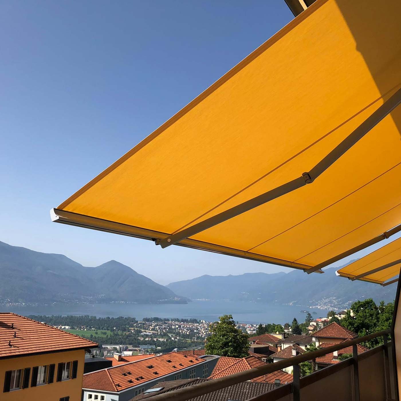 Gelenkarmmarkise mit orangem Tuch von unten, dahinter Dachlandschaft und See