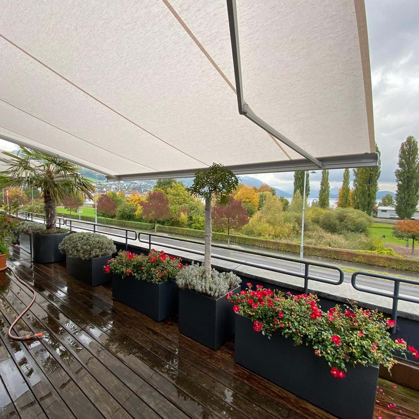 Untersicht einer Markise mit hellem Tuch über einer Holzterrasse mit Pflanzkübeln