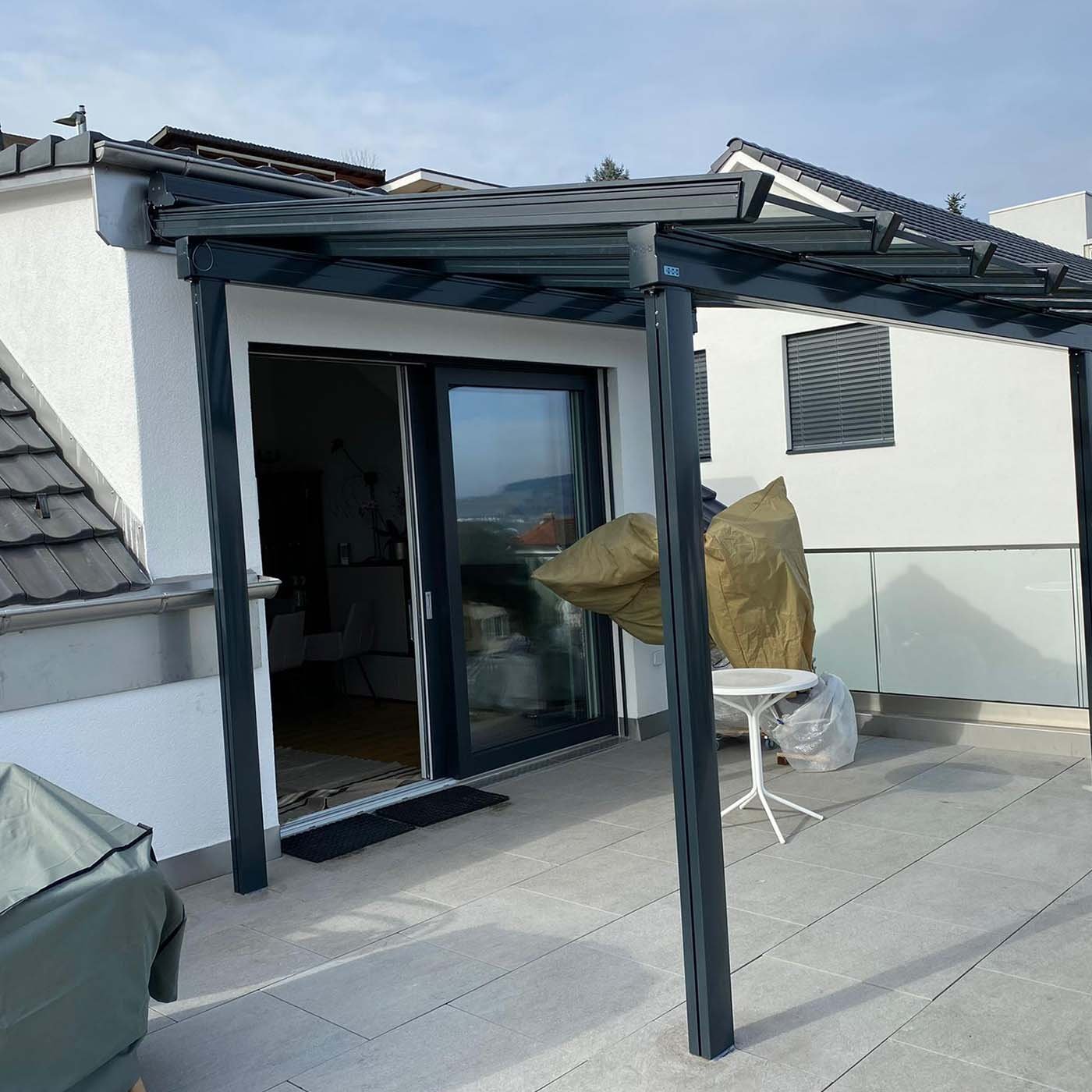 Terrassendach mit Glasdach und anthrazitfarbenem Rahmen an einem Wohnhaus, darunter eine Terrasse mit Glasgeländer