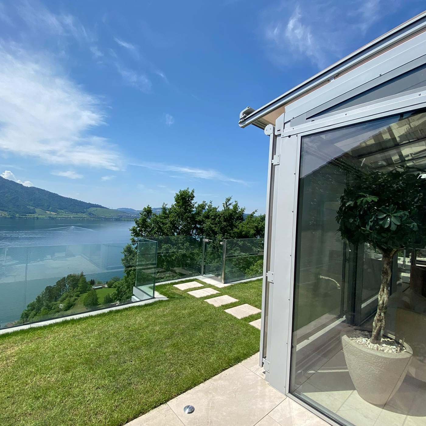 Verglaster Terrassenanbau am Hang, davor ein Glasgeländer und Blick über einen See